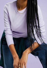 Levandulový pulovr z měkké látky, s kulatým výstřihem, žebrovanými manžetami, v kombinaci s tmavými džínami. Výrazný cop a zlatý řetízek jako doplněk.