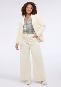 Blazer color crema e pantaloni a gamba larga realizzati in morbido tessuto, abbinati a un top a motivo bianco e nero con dettaglio a chiave.