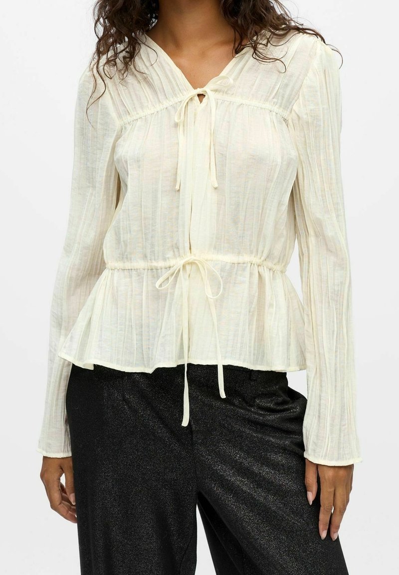 Femme portant un chemisier crème à manches longues avec liens à l'avant et pantalon noir brillant, vue du cou jusqu'aux hanches.