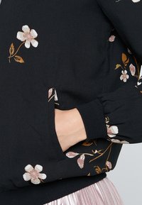 Giacca nera con stampa floreale, realizzata in un tessuto morbido, con maniche a sbuffo e polsini a costine. Include una tasca laterale delineata da un motivo floreale.