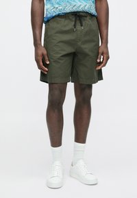 Pantalones cortos caqui con cintura elástica, cordón y bolsillos laterales, combinados con zapatillas blancas y calcetines blancos acanalados.