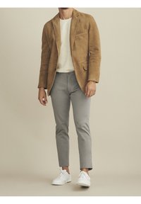 Mies, joka pukeutuu vaaleanruskeaan nahkatakkiin, valkoiseen teksturoituun paitaan, harmaisiin slim-fit-housuihin ja valkoisiin lenkkareihin vaaleanbeigeä taustaa vasten.