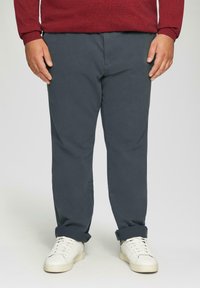 Donkergrijze katoenen chinos met een recht model, opgestikte boorden en zijzakken, gecombineerd met witte sneakers op een effen achtergrond.