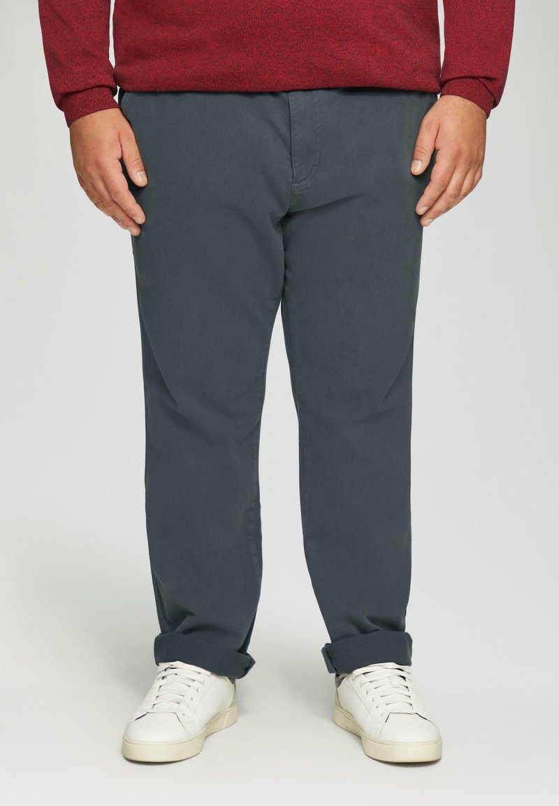 Donkergrijze katoenen chinos met een recht model, opgestikte boorden en zijzakken, gecombineerd met witte sneakers op een effen achtergrond.