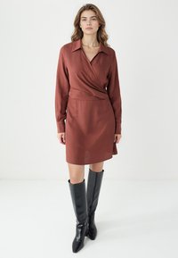 Brązowa sukienka w kroju kopertowym z długimi rękawami, kołnierzykiem i zwężoną talią, zestawiona z czarnymi kozaczkami do kolan. Gładka faktura materiału.