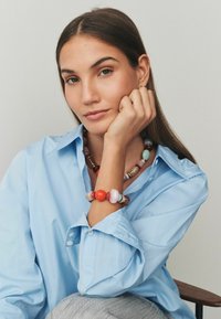 Kaklarota sastāv no daudzkrāsainām pērlēm dažādās formās un izmēros, ieskaitot oranžus, brūnus un pasteļtoņus; valkāta ar gaiši zilu krekls.