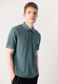 Polo skjorte i en dempet teal farge med et mykt, teksturert stoff. Har klassisk krage, to-knapps placket og subtile logodetaljer.