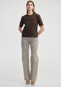 Bruine T-shirt met korte mouwen en gemeleerde branding op de borst, gecombineerd met beige patroon wijdlopende broek en metallic schoenen met een puntige neus.