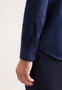 Maglietta a maniche lunghe blu navy con polsini abbottonati, texture elegante e ricamo del logo discreto sulla manica. Vestibilità sagomata mostrata su una mano.