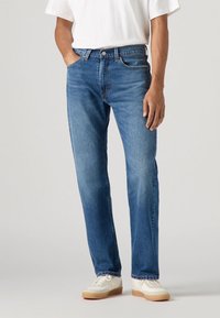 Blauwe spijkerbroeken hebben een rechte snit, een vijf-pocket ontwerp en zichtbare stiksels. Draag ze met een wit T-shirt en beige sneakers.