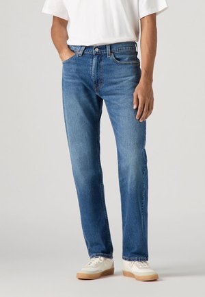 Los jeans de mezclilla azul presentan un corte recto, diseño de cinco bolsillos y costuras visibles. Llevados con una camiseta blanca y zapatillas beige.