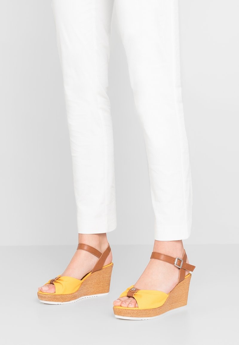 Pieds portant des sandales compensées jaunes avec des brides marron à la cheville, associées à un pantalon blanc court, debout sur un fond clair uni.
