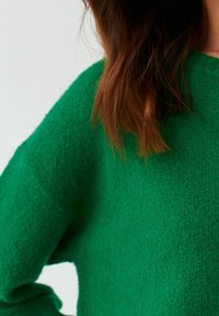 Femme portant un pull vert texturé, aux longs cheveux bruns partiellement visibles, sur un fond blanc uni.