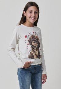 Lächelndes Mädchen, das ein weißes Langarmshirt mit einem Grafikdruck eines Hundes in Sonnenbrille und blaue Jeans mit Strassdetails trägt.