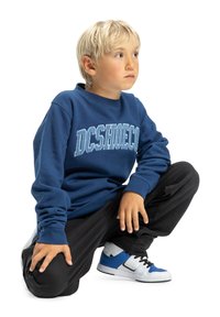 Modrá mikina s logom "DCSHOECO", čierne nohavice a biele tenisky s modrými akcentmi. Dieťa sa skrčené na bielom pozadí.