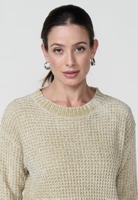 Heled helebežse tekstuuriga kootud sviiter, millel on soonikkoes kaelus, lõtv sobivus ja peen muster kogu ulatuses. Ümmargused hõbedased kõrvarõngad on nähtavad.