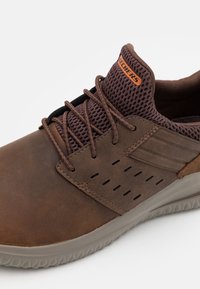 Sneaker in pelle marrone con tomaia in rete, caratterizzata da accenti perforati e lacci neri. Sole in gomma grigia e logo arancione sul tallone.