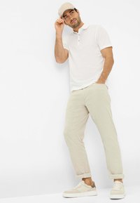 Hvid polo skjorte med to knapper, lys beige linnedbukser og beige sneakers. Kappen er lysebrun med blå akcent.