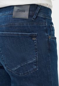 Jeans in denim blu scuro Brax con una toppa in pelle, tasche posteriori con dettagli cuciti e una texture morbida e liscia.