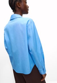 Jasnoniebieska koszula z długim rękawem, z kołnierzykiem, gładką fakturą, zaokrąglonym dołem i zapinanymi mankietami. Noszona na ciemnopomarańczowych spodniach.