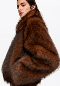 Brązowy płaszcz z futra syntetycznego z szerokim kołnierzem, obszerne rękawy i puszystą fakturę. Posiada ciemne paski wzdłuż materiału.