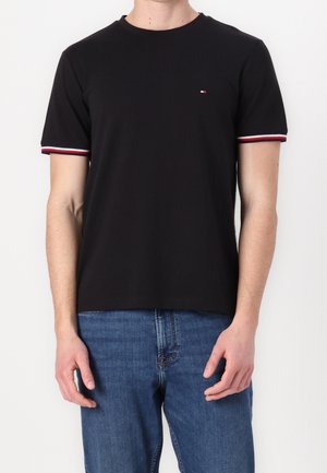 Mand iført en sort t-shirt med korte ærmer med rødt, hvidt og blåt kantbånd på ærmerne og et lille logo på brystet, kombineret med blå jeans.