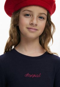 Navyfarbener Ripp-Strickpullover mit pinkfarbener "desigual" Stickerei; das Modell trägt eine rote Filzkappe und präsentiert einen lässigen, schichten Look.