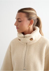 Krémový fleece pulovr s vysokým límcem, zipovým zapínáním a dvěma kulatými hnědými knoflíky. Měkká textura a oversized střih.