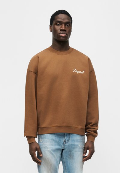 Νεαρός άνδρας φορά καφέ φούτερ με μακριά μανίκια και λευκό λογότυπο "Dsquared2" καθώς και ανοιχτομπλε τζιν με φθορές, στέκεται μπροστά σε ένα απλό φόντο.