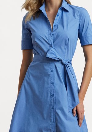 Femme portant une robe chemise bleue courte à manches courtes, longueur genoux, avec col, boutonnage devant et ceinture nouée à la taille.