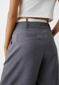 Šedé upravené kalhoty s hladkou texturou, plochými zadními kapsami a bílým pasem. Design je elegantní a přiléhavý v pase.