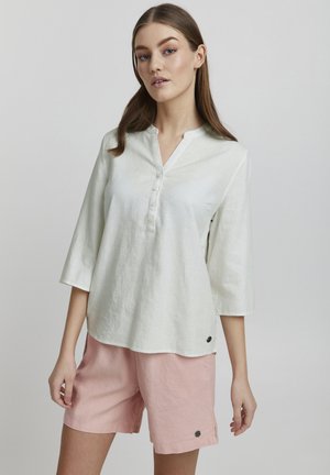 Camisa de lino verde claro con cuello y mangas tres cuartos, con botones en la parte frontal, combinada con pantalones cortos de lino rosa que tienen bolsillos laterales y detalles.