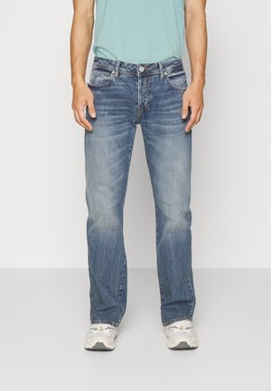 Mann trägt verblasste blaue Jeans mit geradem Bein, hellblaues, in die Hose gestecktes T-Shirt und weiße Turnschuhe, steht vor einem einfarbigen Hintergrund.