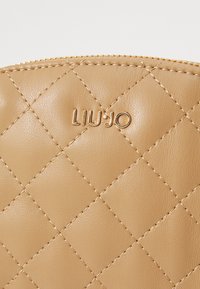 Bolso de piel acolchada beige con el logo dorado "LIU JO", que presenta una superficie texturizada y un cierre de cremallera en la parte superior.