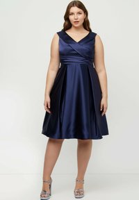 Marinblå satin klänning med V-ringning, korsad midja och plisserad kjol. Kombineras med silverfärgade strappy heels som visar en slät textur.