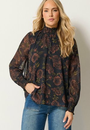 Vrouw met lang blond haar die een zwarte doorschijnende bloes met bloemenprint draagt, met een gesmokte hoge hals en manchetten, gecombineerd met blauwe spijkerbroek, staand tegen een lichte muur.