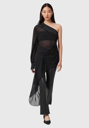 Zwarte asymmetrische top met één lange doorschijnende mouw, gedrapeerd voorkantontwerp, gecombineerd met aansluitende zwarte broek en versierde zwarte schoenen.