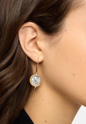 Zbliżenie na ucho z długimi brązowymi włosami, noszące złotą kolczyk w kształcie koła z ozdobną srebrną monetą i małym zwisającym kamieniem szlachetnym.