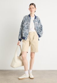 Veste bomber florale dans des tons de bleu, t-shirt blanc, shorts beiges, baskets blanches, tenant un sac fourre-tout crème. Silhouette simple et moderne.