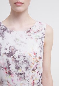 Zbliżenie na osobę ubraną w bezrękawnikową sukienkę w kwiaty o jasnej, pastelowej palecie kolorów na jednolitym tle.