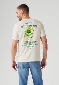 T-shirt crème avec un grand graphique de marguerite jaune au dos et du texte vert. Coupe décontractée, manches courtes, présenté sur un fond neutre.