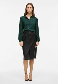 Camicia button-up verde con texture e maniche lunghe, abbinata a una gonna a matita nera con spacco frontale, indossata con sandali neri con tacco.