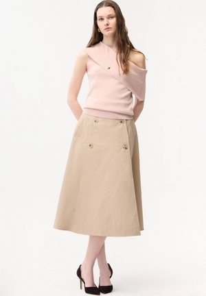Pitkähiuksinen nainen, joka pukeutunut vaaleanpunaiseen epäsymmetriseen hihattomaan toppiin, beigenväriseen nappilliseen midihameeseen ja mustiin korkeakorkoisiin kenkiin, seisoo valkoista taustaa vasten.