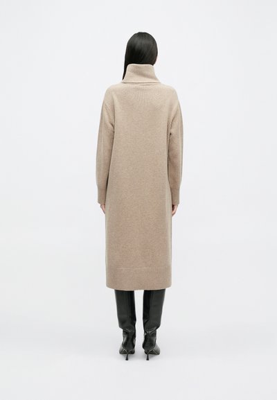 Duga bež pletena haljina s visokim ovratnikom, rebrastim manšetama i donjim rubom. Nošena preko crnih uskih čizama do koljena.