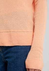 Persikofärgad stickad tröja med ribbad fåll och muddar, med ett diskret hålmönster längs nederkanten. Buren över blå jeans i denim.