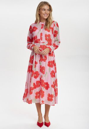 Blommig midiklänning i genomskinlig rosa tyg med röda vallmo-blommor, långa ärmar, knappstängning framtill och designer i lagerkjol. Röda spetsiga klackskor.