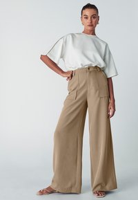 Maglietta bianca a maniche corte con decorazioni dorate, abbinata a pantaloni beige a vita alta e gamba larga con tasche frontali e una texture liscia.