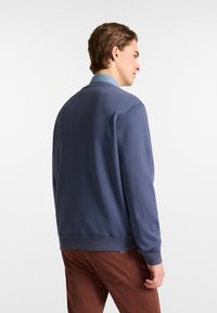 Marineblaues Sweatshirt mit Rundhalsausschnitt, gerippten Bündchen und Saum, getragen über einem hellblauen Hemd mit Kragen, kombiniert mit braunen Hosen.