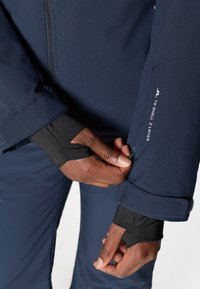 Chaqueta de dos capas TX-Shell azul marino con cremallera, que presenta detalles en negro en los puños y un acabado en las costuras. Primer plano de la manga ajustable con el logo.