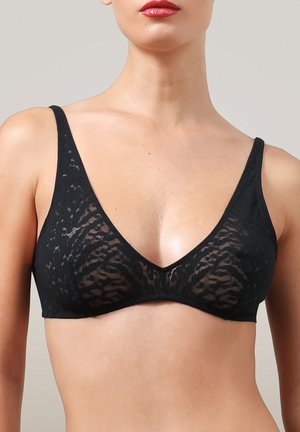 Vrouw draagt een zwarte kanten draadloze beha met dunne straps en een diepe V-halslijn tegen een effen achtergrond.